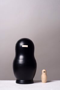 Fünfteilige Matrjoschka-Puppe, Kunstwerk „o.T.“ (2013) von Simone Brühl. Die Puppen sind nach Größe sortiert aufgereiht. Die vier größeren Figuren sind schwarz besprüht und haben eine rechteckige Holzaussparung im Gesicht. Die kleinste, innerste Figur ist aus hellem Naturholz und hat stattdessen einen schwarzen Balken im Gesicht.