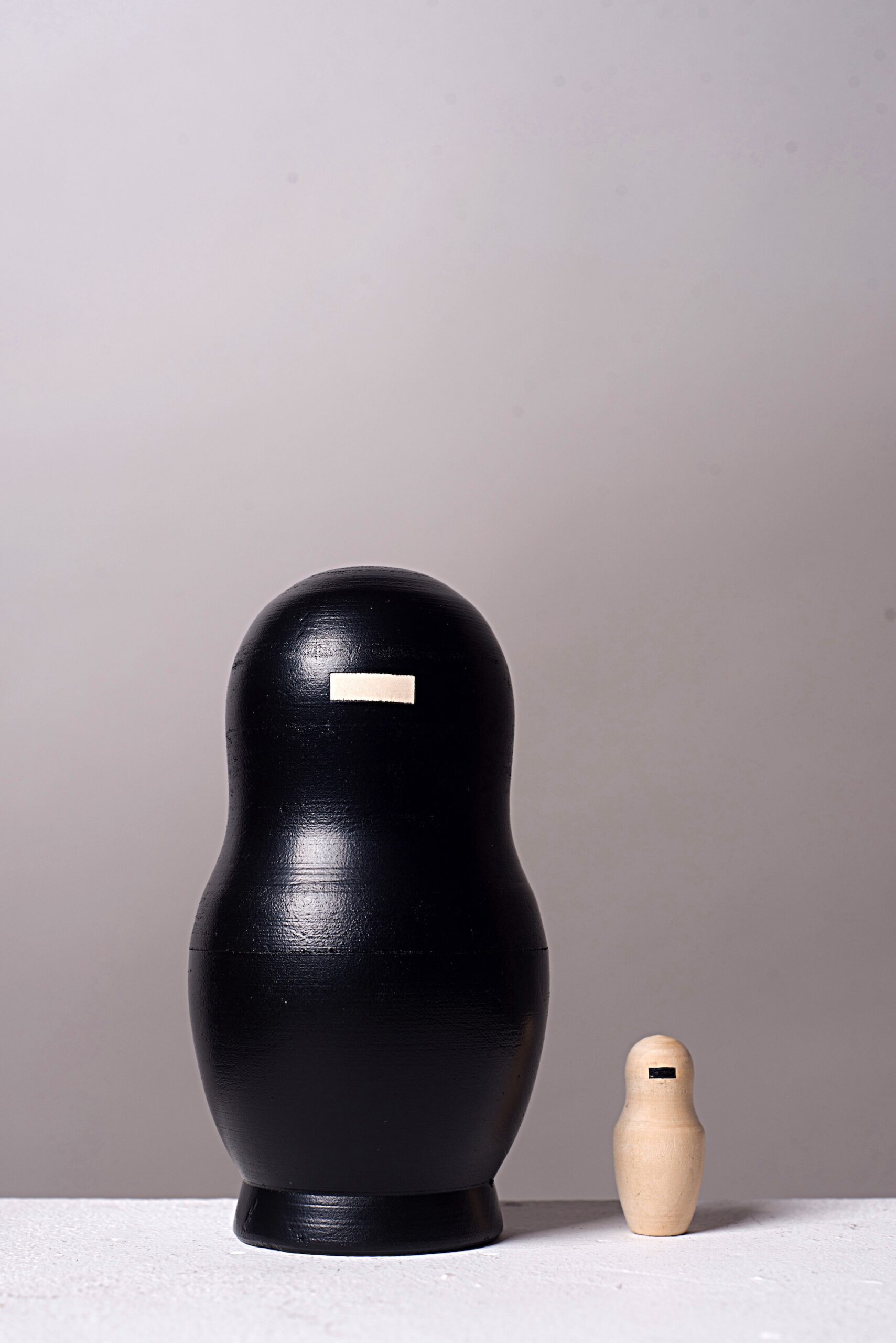 Fünfteilige Matrjoschka-Puppe, Kunstwerk „o.T.“ (2013) von Simone Brühl. Die Puppen sind nach Größe sortiert aufgereiht. Die vier größeren Figuren sind schwarz besprüht und haben eine rechteckige Holzaussparung im Gesicht. Die kleinste, innerste Figur ist aus hellem Naturholz und hat stattdessen einen schwarzen Balken im Gesicht.