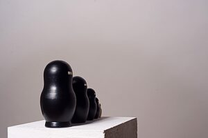 Fünfteilige Matrjoschka-Puppe, Kunstwerk „o.T.“ (2013) von Simone Brühl. Die Puppen sind nach Größe sortiert aufgereiht. Die vier größeren Figuren sind schwarz besprüht und haben eine rechteckige Holzaussparung im Gesicht. Die kleinste, innerste Figur ist aus hellem Naturholz und hat stattdessen einen schwarzen Balken im Gesicht.