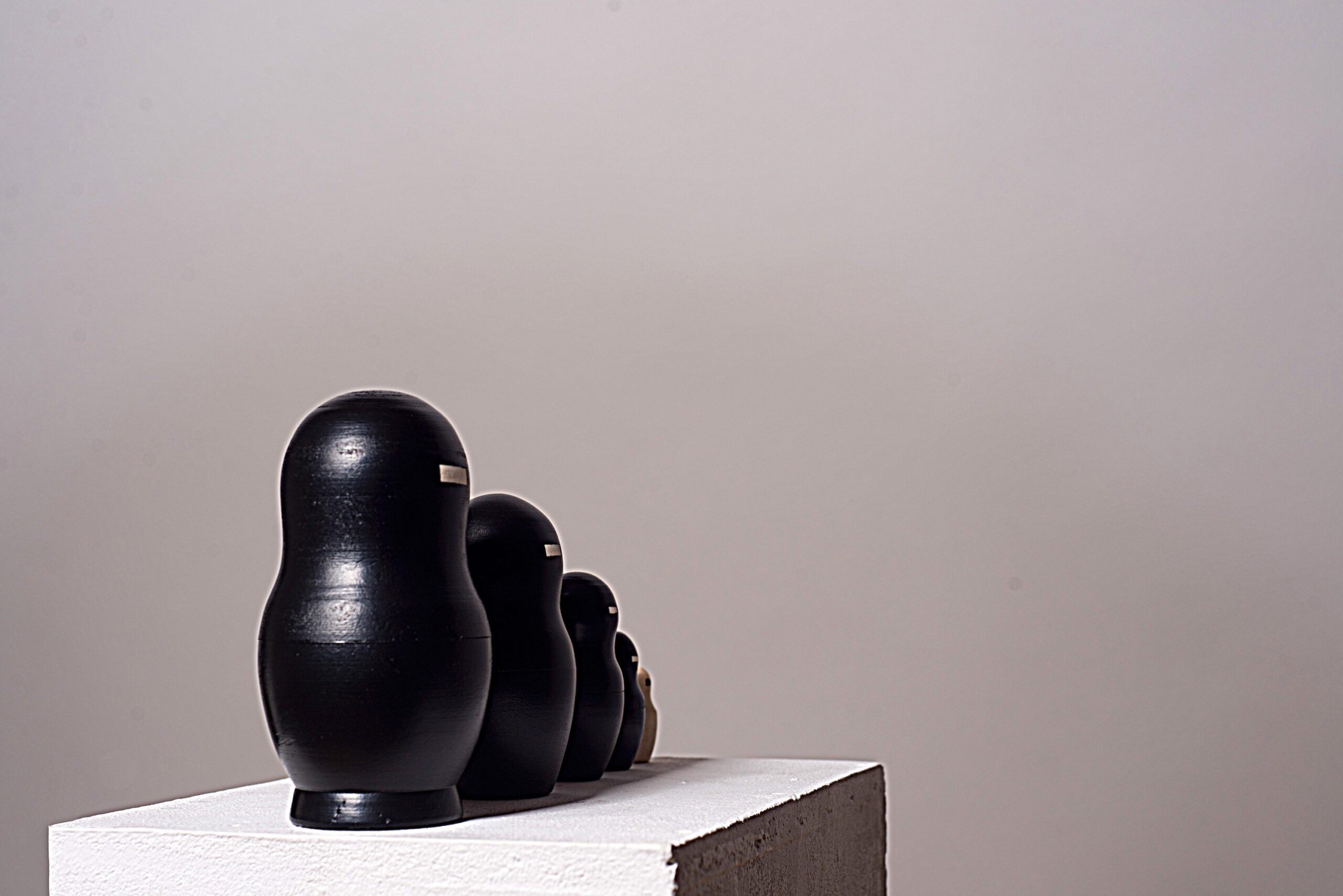 Fünfteilige Matrjoschka-Puppe, Kunstwerk „o.T.“ (2013) von Simone Brühl. Die Puppen sind nach Größe sortiert aufgereiht. Die vier größeren Figuren sind schwarz besprüht und haben eine rechteckige Holzaussparung im Gesicht. Die kleinste, innerste Figur ist aus hellem Naturholz und hat stattdessen einen schwarzen Balken im Gesicht.