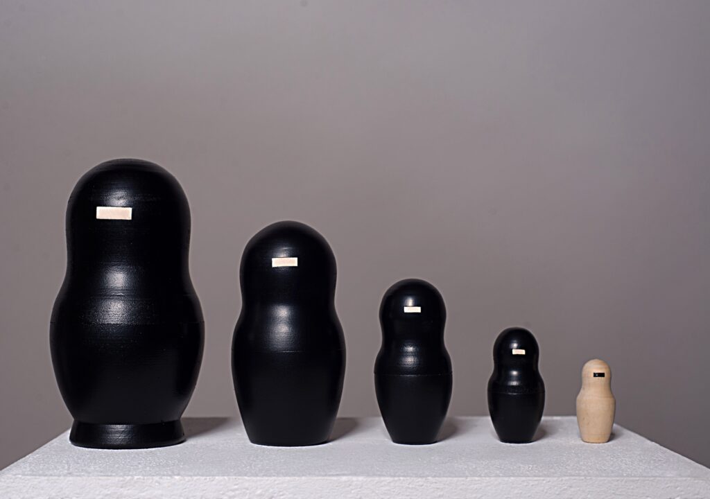 Das 2013 entstandene, fünfteilige Kunstwerk „o.T.“ von Simone Brühl nutzt die Struktur einer traditionellen 
Matrjoschka-Puppe, um durch gezielte Reduktion die Themen Identität, Schichtung und Anonymisierung zu untersuchen.

Durch den Einsatz von schwarzem Sprühlack auf den vier größeren, äußeren Figuren werden deren übliche, individuelle Bemalungen vollständig überdeckt, was den Fokus auf die reine Grundform lenkt. Das zentrale Gestaltungsmerkmal basiert dabei auf dem Prinzip der Farbinversion: Die vier schwarzen Hüllen weisen im Gesichtsbereich eine rechteckige Aussparung auf, die das helle Naturholz der Puppen freigibt. Bei der kleinsten, innersten Figur verhält es sich umgekehrt – sie verbleibt im natürlichen Holzton, ist jedoch im selben Bereich mit einem schwarzen Farbbalken versehen.

Diese formale Umkehrung und die monochrome Übermalung erzeugen ein Wechselspiel, das die visuelle und konzeptuelle Trennung zwischen den umschließenden äußeren Schichten und dem verborgenen, inneren Kern der Objekte thematisiert.