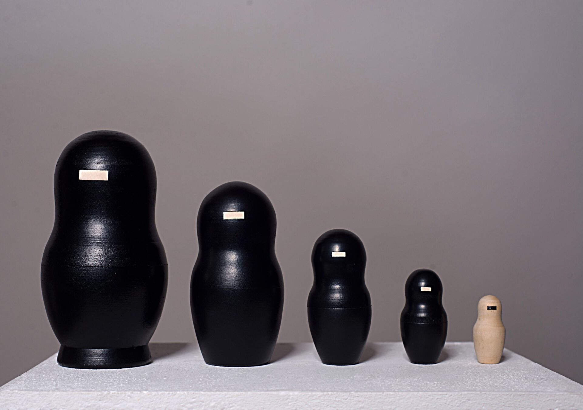 Fünfteilige Matrjoschka-Puppe, Kunstwerk „o.T.“ (2013) von Simone Brühl. Die Puppen sind nach Größe sortiert aufgereiht. Die vier größeren Figuren sind schwarz besprüht und haben eine rechteckige Holzaussparung im Gesicht. Die kleinste, innerste Figur ist aus hellem Naturholz und hat stattdessen einen schwarzen Balken im Gesicht.