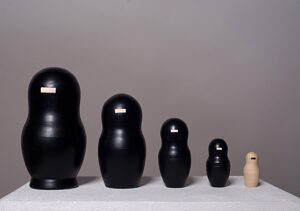 Fünfteilige Matrjoschka-Puppe, Kunstwerk „o.T.“ (2013) von Simone Brühl. Die Puppen sind nach Größe sortiert aufgereiht. Die vier größeren Figuren sind schwarz besprüht und haben eine rechteckige Holzaussparung im Gesicht. Die kleinste, innerste Figur ist aus hellem Naturholz und hat stattdessen einen schwarzen Balken im Gesicht.